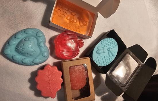 “Handmade soaps in various shapes and colors, including pumpkins, leaves, hearts, and honeycomb designs, displayed in gift-ready boxes with clear windows.”
